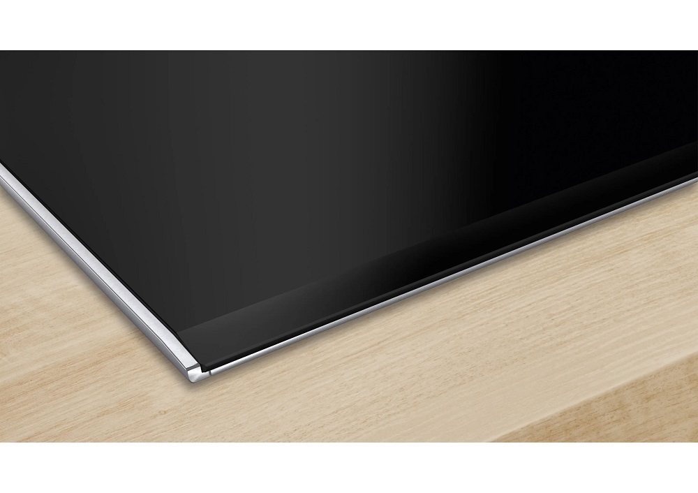 Macro view of the flat ceramic glass edge of the cooktop with a slim stainless steel trim on a wooden countertop.