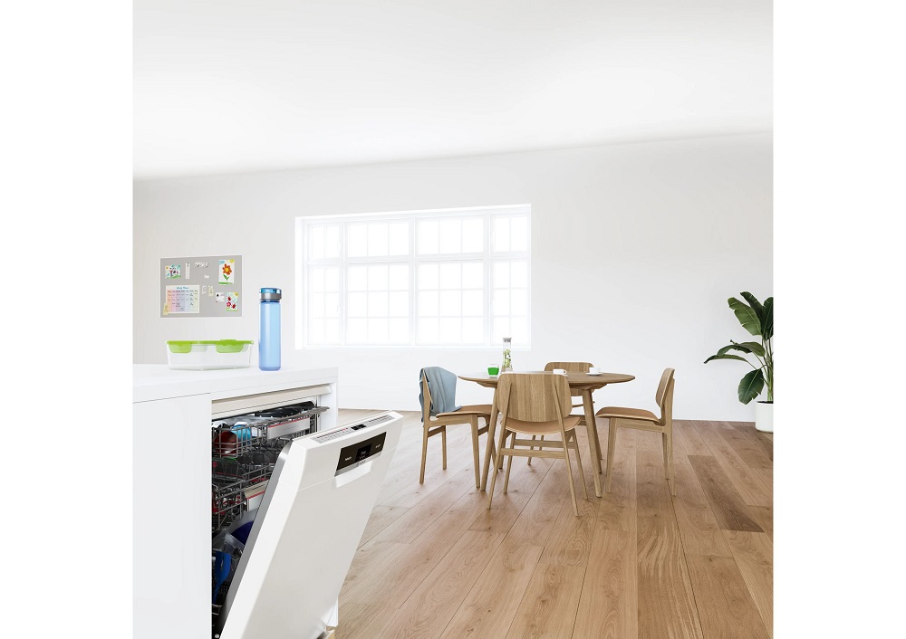 Brightly lit lifestyle shot of a modern, minimalist kitchen looking towards a dining area, featuring a partially opened, fully integrated Bosch dishwasher installed in a white kitchen island in the foreground.