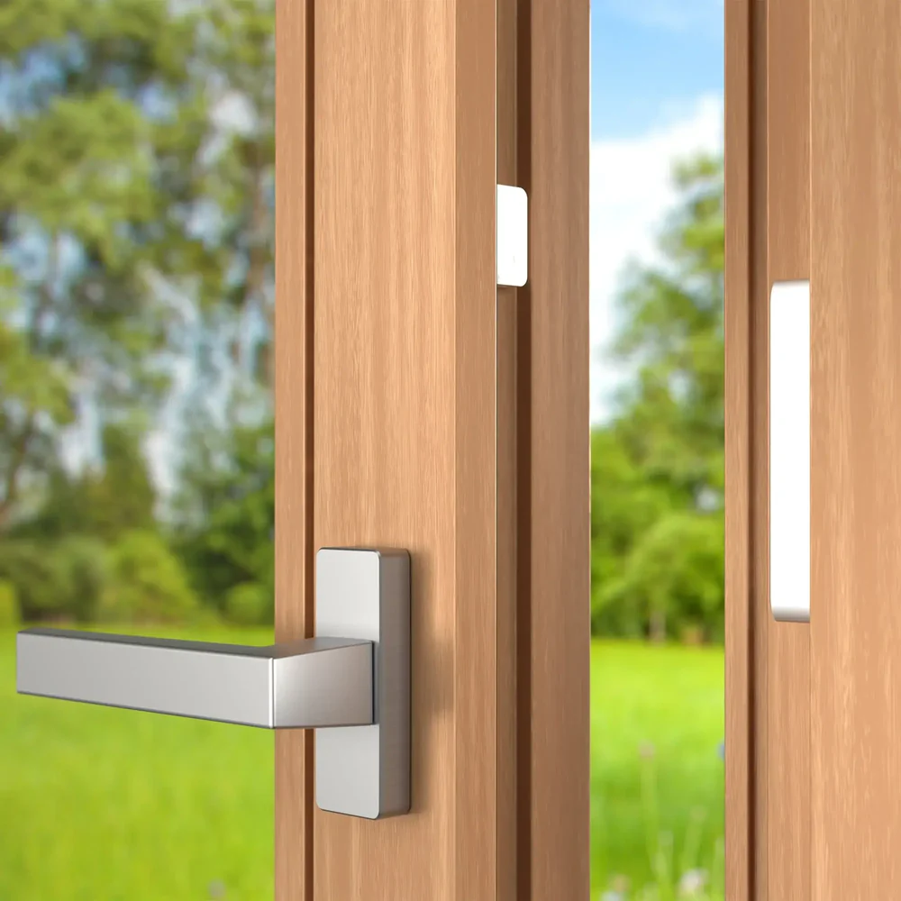 Der magnetische Fenstersensor (kleiner und großer Teil) ist diskret an einem geöffneten Holzfenster montiert, mit grünem Garten im Hintergrund.