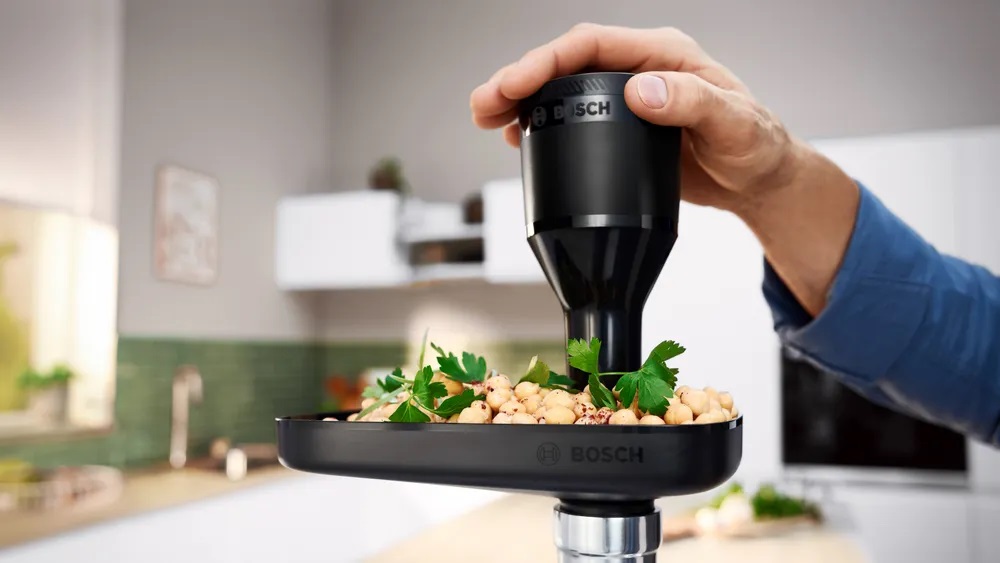 Der Bosch MFWS420W Kompakt-Fleischwolf verarbeitet Kichererbsen und Kräuter in seinem Einfülltrichter, demonstriert seine Vielseitigkeit für vegetarische Gerichte.
